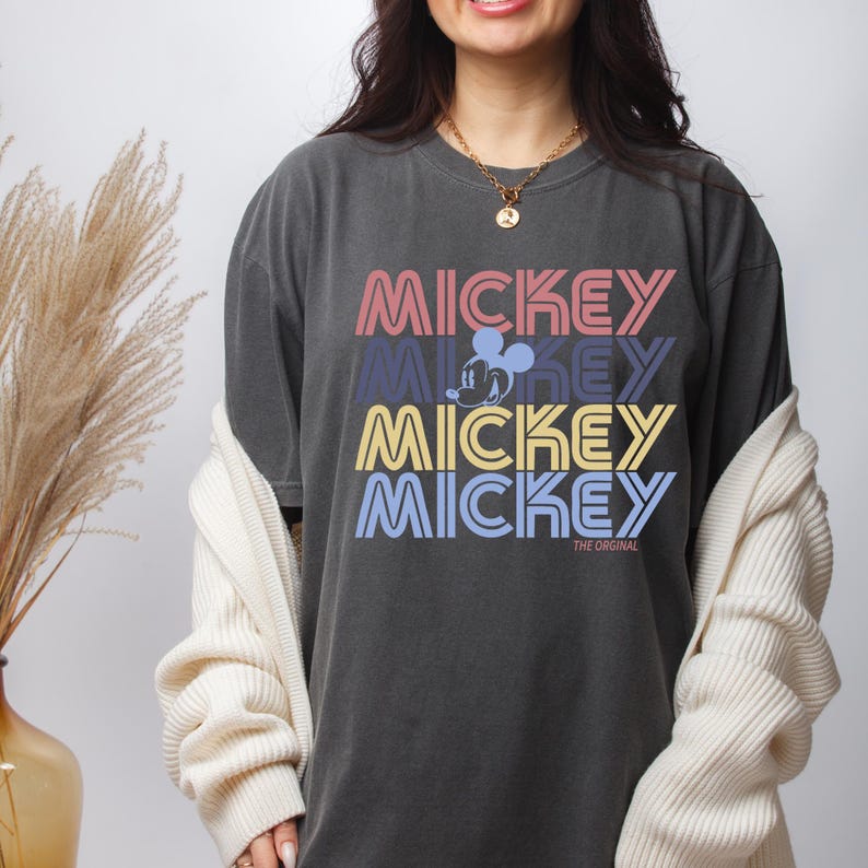 May include: Dark gray t-shirt with the word "MICKEY" in pink, purple, yellow, and blue. The shirt features a Mickey Mouse graphic and the text "THE ORIGINAL". The shirt is part of a Disney-themed collection.