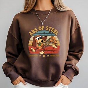 Puede incluir: Una sudadera marr&oacute;n con un dise&ntilde;o de perro salchicha de dibujos animados. El dise&ntilde;o incluye el texto "ABS OF STEEL" y una puesta de sol retro en rojo, naranja, amarillo y azul. La sudadera tiene cuello redondo y mangas largas.