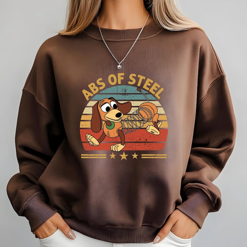 Puede incluir: Una sudadera marr&oacute;n con un dise&ntilde;o de perro salchicha de dibujos animados. El dise&ntilde;o incluye el texto "ABS OF STEEL" y una puesta de sol retro en rojo, naranja, amarillo y azul. La sudadera tiene cuello redondo y mangas largas.