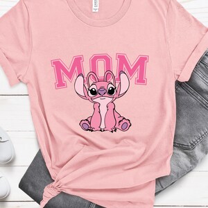 May include: A light pink short-sleeved t-shirt featuring a graphic of the character Angel from Lilo and Stitch sitting down. Above the character, the word MOM is printed in a bold, dark pink collegiate-style font with a lighter pink outline.