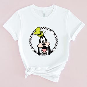 Op de afbeelding: Wit T-shirt met een zwart-wit geblokte cirkel rond een cartoon afbeelding van Goofy. Goofy draagt een gele hoed en heeft een brede glimlach.