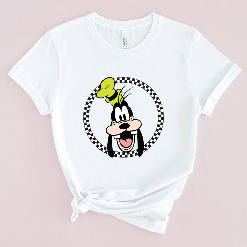 Op de afbeelding: Wit T-shirt met een zwart-wit geblokte cirkel rond een cartoon afbeelding van Goofy. Goofy draagt een gele hoed en heeft een brede glimlach.