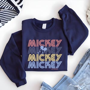 May include: Navy blue sweatshirt featuring the word "MICKEY" stacked in retro colors: coral, gray, yellow, and blue. A Mickey Mouse graphic is centered between the words. The text "THE ORIGINAL" is printed below the bottom word. The sweatshirt is folded on a white surface.