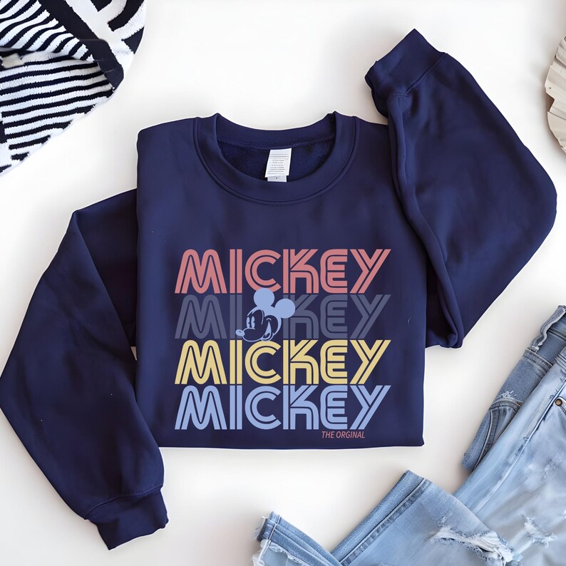 May include: Navy blue sweatshirt featuring the word "MICKEY" stacked in retro colors: coral, gray, yellow, and blue. A Mickey Mouse graphic is centered between the words. The text "THE ORIGINAL" is printed below the bottom word. The sweatshirt is folded on a white surface.
