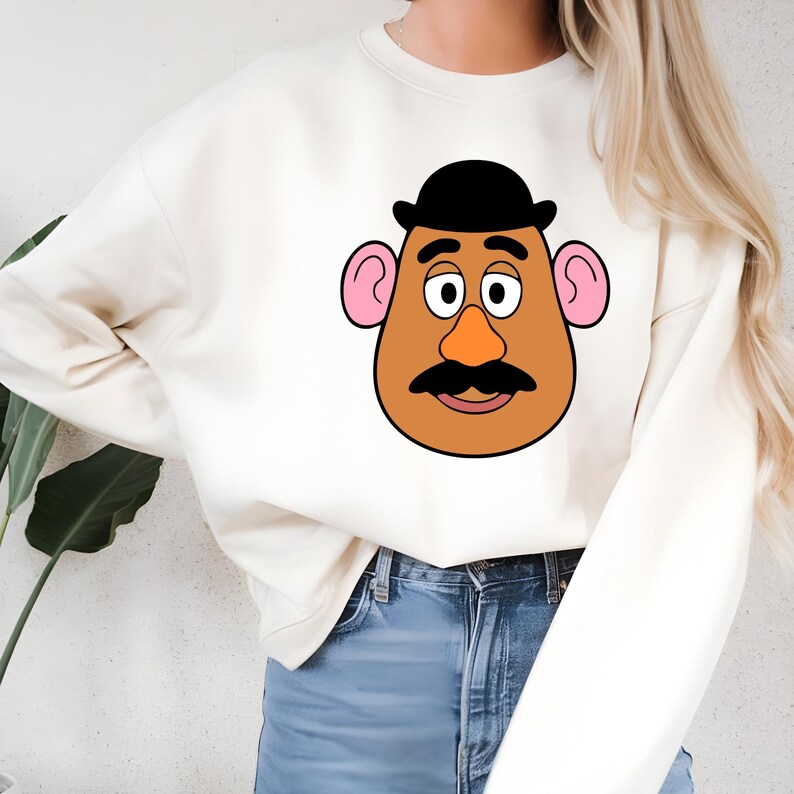 May include: Cream-colored sweatshirt featuring a cartoon illustration of a potato head character. The character has a brown face, black mustache, black bowler hat, pink ears, and an orange nose. The sweatshirt is a casual style.
