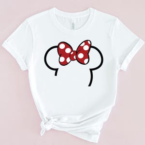 Op de afbeelding: Wit T-shirt met een rode strik met witte stippen en zwarte muizenoren. Het ontwerp staat in het midden van de voorkant van de shirt. Het shirt heeft korte mouwen en een ronde hals.