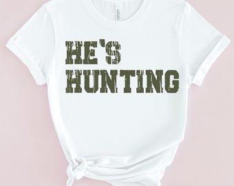 May include: White t-shirt with the text "HE'S HUNTING" in green distressed lettering.