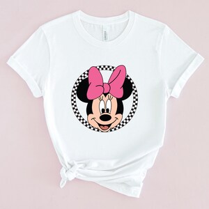 Op de afbeelding: Wit T-shirt met een zwart-wit geblokte cirkel rond een cartoon afbeelding van Minnie Mouse met een roze strik.