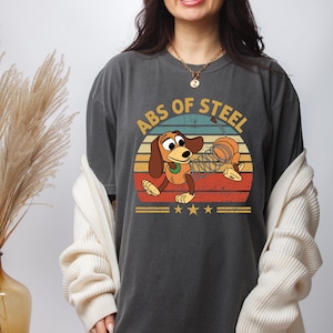 Puede incluir: Camiseta gris oscuro con un dise&ntilde;o de perro de dibujos animados con un cuerpo de resorte, sobre un fondo de puesta de sol retro. El texto "ABS OF STEEL" est&aacute; encima del perro. Debajo hay tres estrellas doradas y dos l&iacute;neas horizontales.