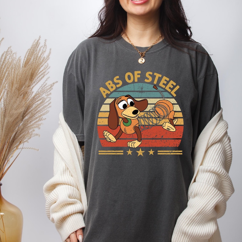 Puede incluir: Camiseta gris oscuro con un dise&ntilde;o de perro de dibujos animados con un cuerpo de resorte, sobre un fondo de puesta de sol retro. El texto "ABS OF STEEL" est&aacute; encima del perro. Debajo hay tres estrellas doradas y dos l&iacute;neas horizontales.