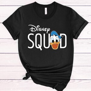 Puede incluir: Camiseta negra con el logo de Disney y la palabra "SQUAD" en blanco, junto con una ilustraci&oacute;n de dibujos animados de Pato Donald. La camiseta tiene mangas cortas y cuello redondo.