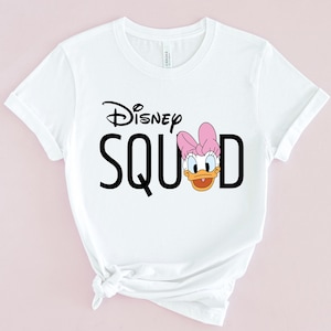 Puede incluir: Camiseta blanca con la frase "Disney SQUAD" en letras negras. La letra "D" es reemplazada por una imagen de dibujos animados de Daisy Duck con un lazo rosa. La camiseta est&aacute; hecha de un material suave.