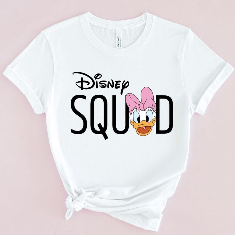 Puede incluir: Camiseta blanca con la frase "Disney SQUAD" en letras negras. La letra "D" es reemplazada por una imagen de dibujos animados de Daisy Duck con un lazo rosa. La camiseta est&aacute; hecha de un material suave.