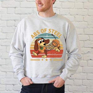 Puede incluir: Sudadera gris claro con un dise&ntilde;o de perro salchicha de dibujos animados. El perro tiene un cuerpo de resorte y est&aacute; colocado contra un dise&ntilde;o de puesta de sol retro. El texto "ABS OF STEEL" est&aacute; encima del perro. El dise&ntilde;o incluye estrellas y rayas.