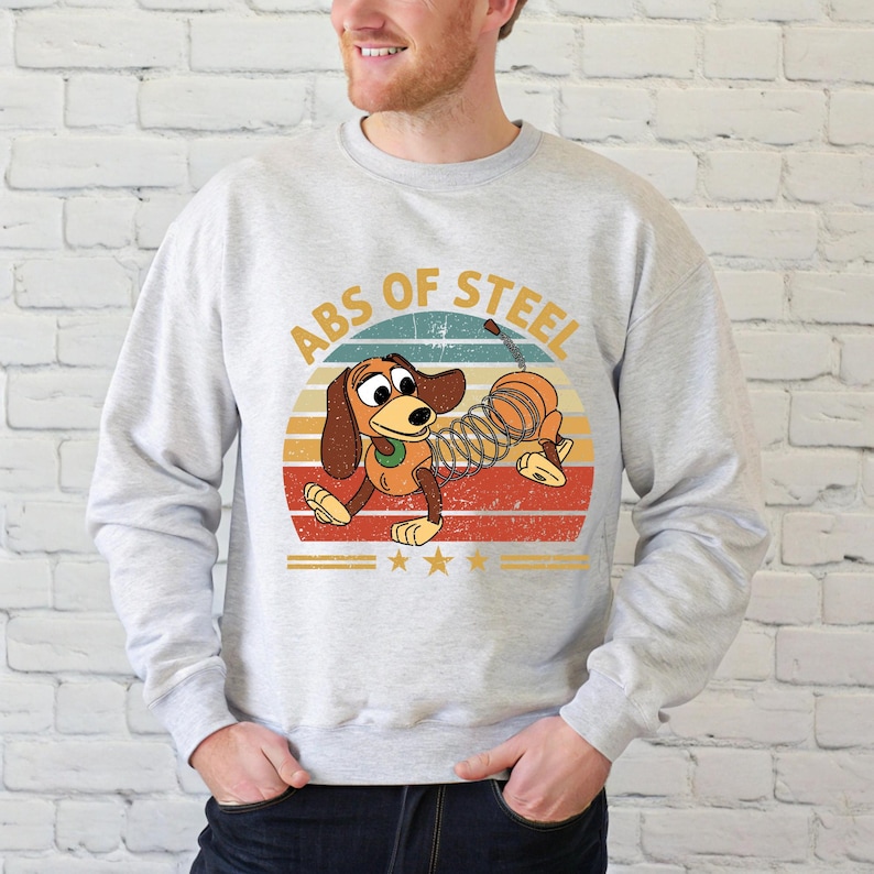 Puede incluir: Sudadera gris claro con un dise&ntilde;o de perro salchicha de dibujos animados. El perro tiene un cuerpo de resorte y est&aacute; colocado contra un dise&ntilde;o de puesta de sol retro. El texto "ABS OF STEEL" est&aacute; encima del perro. El dise&ntilde;o incluye estrellas y rayas.