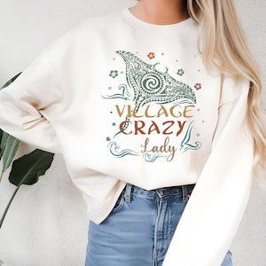 May include: Cream-colored sweatshirt with a graphic design featuring a stylized manta ray and the words "Village Crazy Lady." The design incorporates shades of green, brown, and red, with floral and wave-like accents. The sweatshirt is a casual, comfortable garment.
