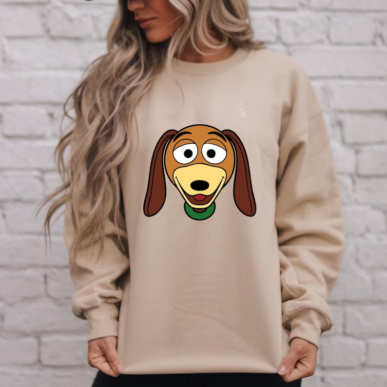 May include: A tan sweatshirt featuring a cartoon image of Slinky Dog from Toy Story. The graphic shows Slinky's face with brown ears, a yellow muzzle, and a green collar. The sweatshirt is a neutral color.