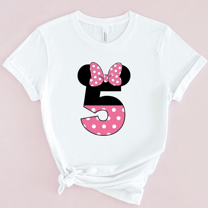 May include: White t-shirt featuring a black number 5 with a pink polka-dotted design. The number is topped with black mouse ears and a pink polka-dotted bow. The shirt is made of a soft material.