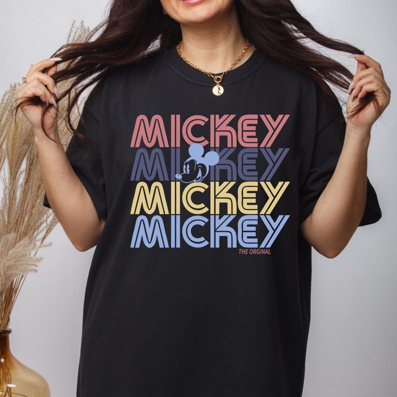 May include: Black t-shirt with the word "MICKEY" in a stacked design, using pink, blue, yellow, and purple colors. The shirt includes a graphic of Mickey Mouse and the text "THE ORIGINAL".