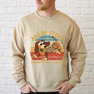 Puede incluir: Sudadera beige con un perro de dibujos animados con cuerpo de resorte, sobre un dise&ntilde;o retro de puesta de sol. El texto "ABS OF STEEL" est&aacute; arqueado sobre el perro. Tres estrellas doradas est&aacute;n debajo.