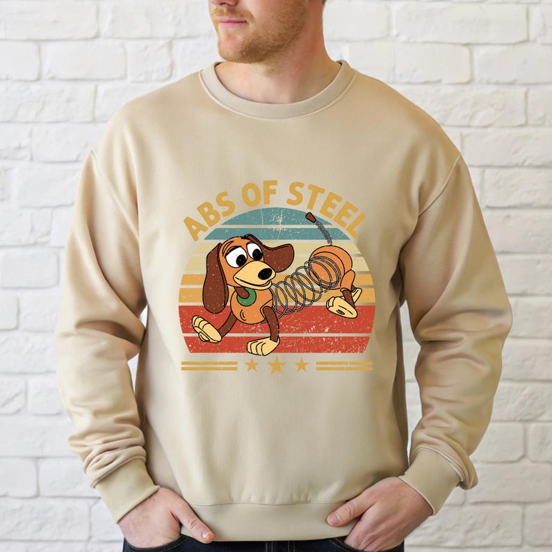 Puede incluir: Sudadera beige con un perro de dibujos animados con cuerpo de resorte, sobre un dise&ntilde;o retro de puesta de sol. El texto "ABS OF STEEL" est&aacute; arqueado sobre el perro. Tres estrellas doradas est&aacute;n debajo.