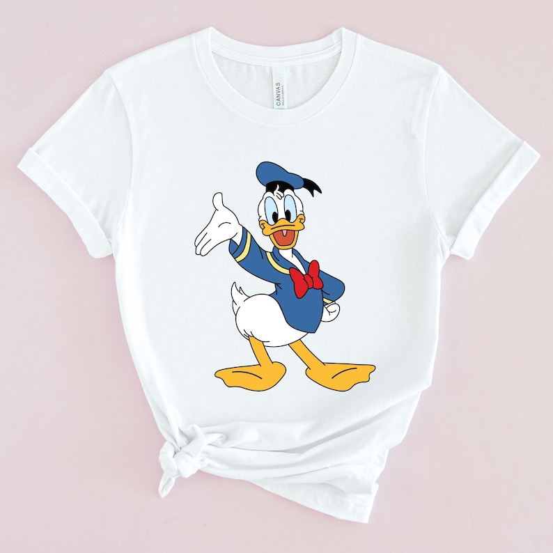 May include: White t-shirt with a cartoon image of Donald Duck wearing a blue sailor shirt and a blue sailor hat. Donald Duck is waving with his right hand and has a red bow tie.