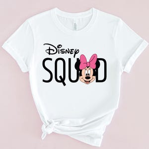 Puede incluir: Camiseta blanca con la frase "Disney Squad" en negro y un dibujo de Minnie Mouse. Minnie Mouse lleva un lazo rosa. La camiseta tiene las mangas enrolladas y cuello redondo.