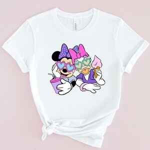 May include: White t-shirt with a graphic of Minnie Mouse and Daisy Duck wearing heart-shaped sunglasses. Minnie is holding a purple drink and Daisy is holding an ice cream cone.