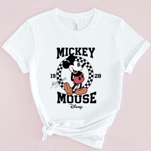 May include: White t-shirt with a black and white graphic of Mickey Mouse in a checkered circle. The text "Mickey Mouse" is above the graphic and "Disney" is below. The text "1928" is on the right side of the circle and "Mickey Mouse" is written in cursive on the left side of the circle.