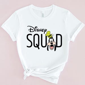 Puede incluir: Camiseta blanca con la frase "Disney SQUAD" en negro, con un dibujo de Goofy. El logo de Disney est&aacute; encima de la palabra "SQUAD". Goofy lleva su sombrero caracter&iacute;stico y tiene una gran sonrisa.