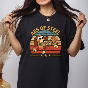 Puede incluir: Camiseta negra con un perro de dibujos animados y el texto "ABS OF STEEL" en un dise&ntilde;o retro. El perro es marr&oacute;n y canela, con un cuerpo en forma de muelle, sobre un fondo de colores de puesta de sol. Tres estrellas doradas debajo.