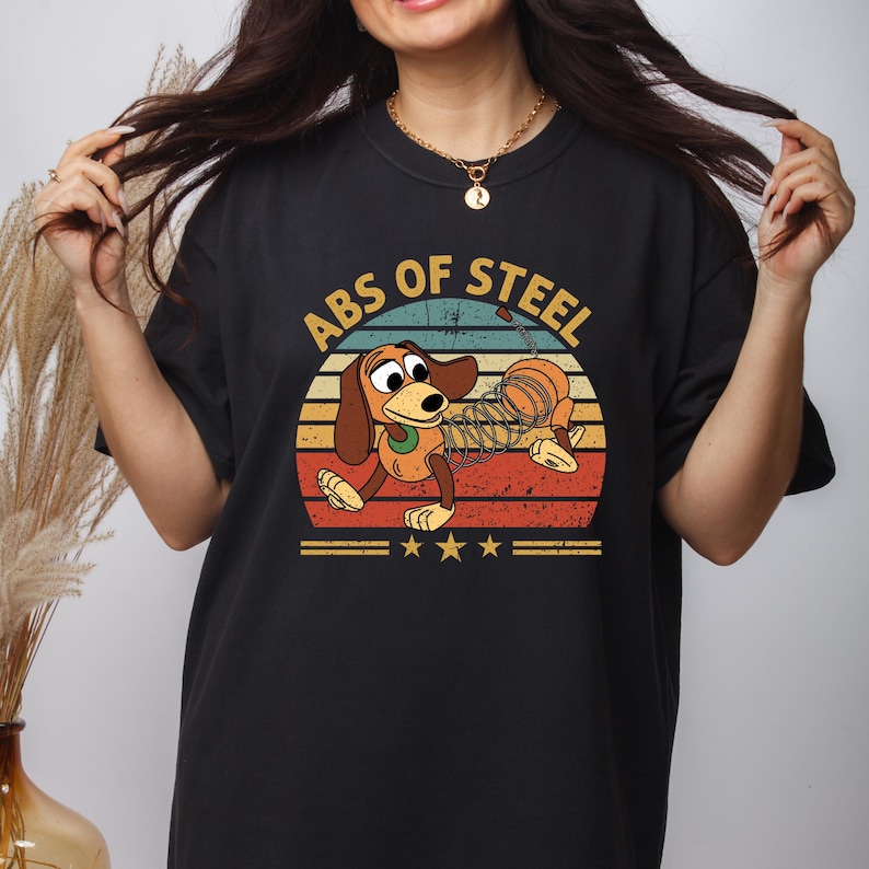 Puede incluir: Camiseta negra con un perro de dibujos animados y el texto "ABS OF STEEL" en un dise&ntilde;o retro. El perro es marr&oacute;n y canela, con un cuerpo en forma de muelle, sobre un fondo de colores de puesta de sol. Tres estrellas doradas debajo.