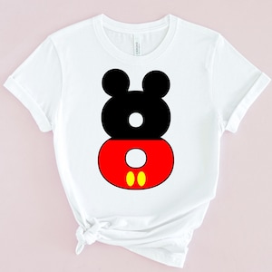 May include: White t-shirt featuring a graphic design of Mickey Mouse's head and shorts. The design is composed of black, red, and yellow shapes. The shirt has short sleeves and a crew neck.
