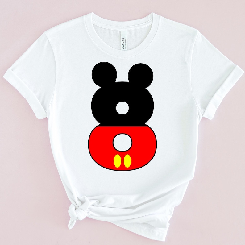 May include: White t-shirt featuring a graphic design of Mickey Mouse's head and shorts. The design is composed of black, red, and yellow shapes. The shirt has short sleeves and a crew neck.