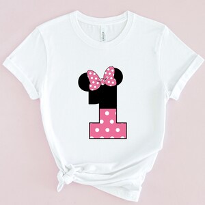 May include: White t-shirt featuring a black number one with a pink polka-dot design. The number one has black mouse ears and a pink polka-dot bow. The shirt is a crew neck and has short sleeves.
