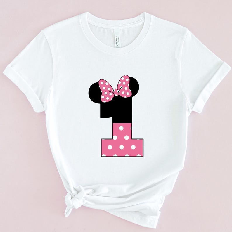 May include: White t-shirt featuring a black number one with a pink polka-dot design. The number one has black mouse ears and a pink polka-dot bow. The shirt is a crew neck and has short sleeves.