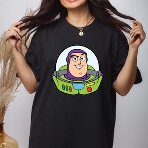May include: Black t-shirt featuring a cartoon graphic of Buzz Lightyear's face. The graphic includes a light blue background, a green space suit, and the character's face in shades of purple, tan, and white. The shirt has short sleeves and a crew neck.