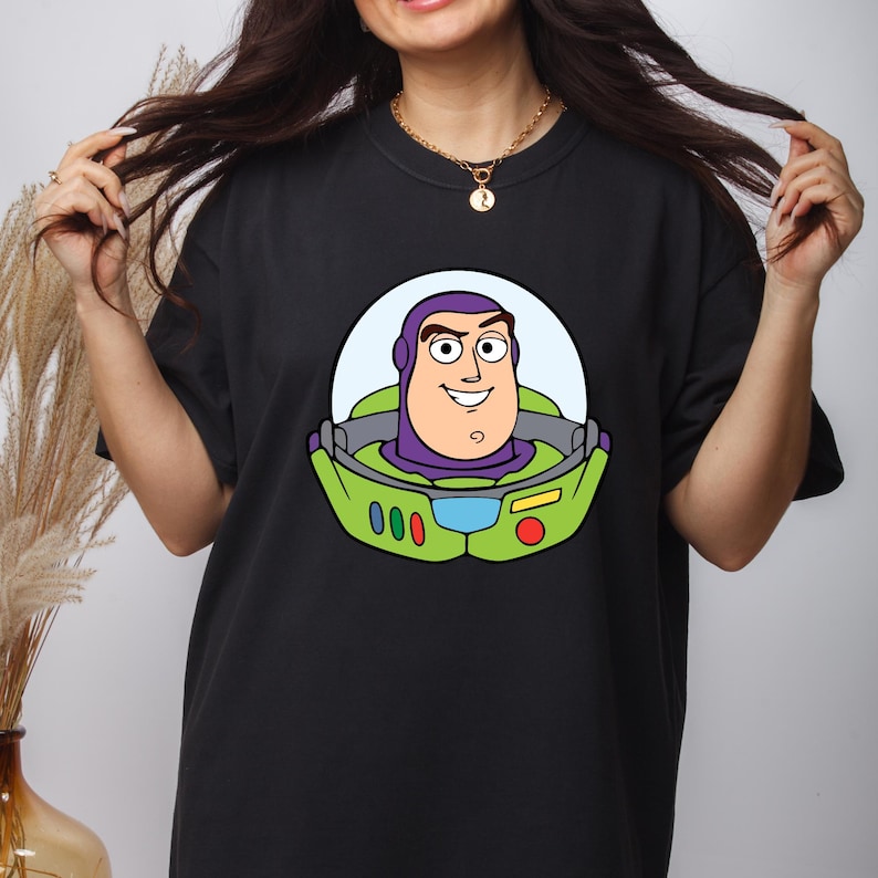 May include: Black t-shirt featuring a cartoon graphic of Buzz Lightyear's face. The graphic includes a light blue background, a green space suit, and the character's face in shades of purple, tan, and white. The shirt has short sleeves and a crew neck.