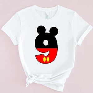 May include: White t-shirt featuring a large number nine in a Mickey Mouse-inspired design. The top portion of the number is black with two black circles, the bottom is red with two yellow circles.