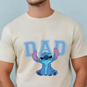 May include: A cream-colored short-sleeve t-shirt featuring the word DAD in large, light blue block letters. Below the text is a graphic illustration of the blue alien character Stitch, who has large ears with pink inner details and a small smile.