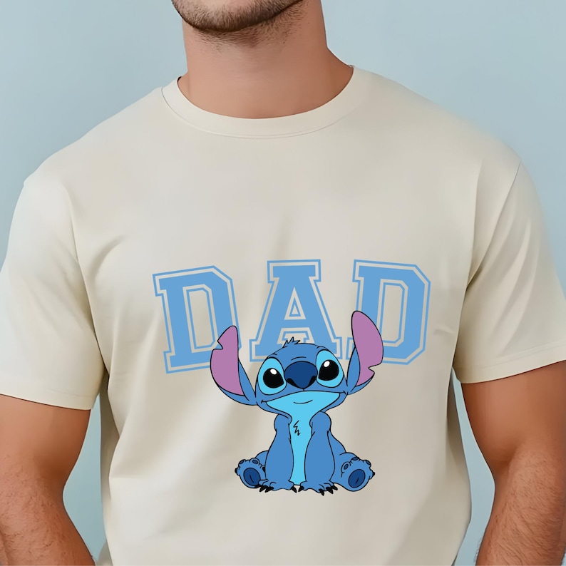 May include: A cream-colored short-sleeve t-shirt featuring the word DAD in large, light blue block letters. Below the text is a graphic illustration of the blue alien character Stitch, who has large ears with pink inner details and a small smile.