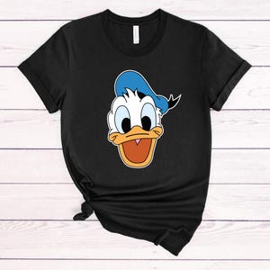 Peut inclure: T-shirt noir avec une image de dessin anim&eacute; du visage de Donald Duck. Donald Duck porte un chapeau de marin bleu.