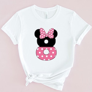 May include: White t-shirt featuring a black and pink graphic of a Minnie Mouse-inspired design. The design includes a black head with a pink polka dot bow, a black number 8, and a pink polka dot circle.