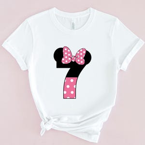May include: White t-shirt featuring a large number 7 design. The number is black with a pink polka dot bow and a pink polka dot pattern on the bottom half. The shirt is made of a soft material.