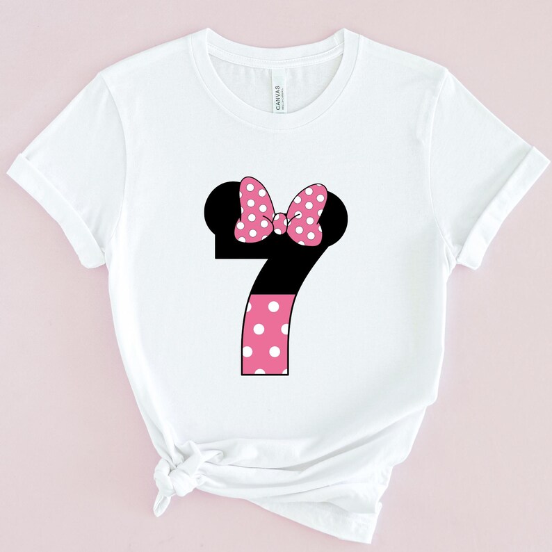May include: White t-shirt featuring a large number 7 design. The number is black with a pink polka dot bow and a pink polka dot pattern on the bottom half. The shirt is made of a soft material.