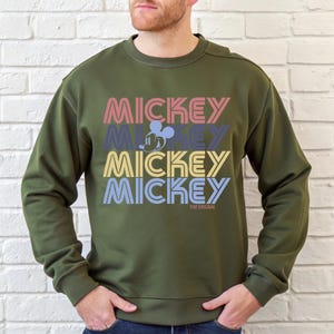 May include: Olive green sweatshirt with the word "MICKEY" stacked four times in different colors. The word "MICKEY" is in pink, blue, yellow, and blue. A Mickey Mouse graphic is in the center. The text "THE ORIGINAL" is at the bottom.