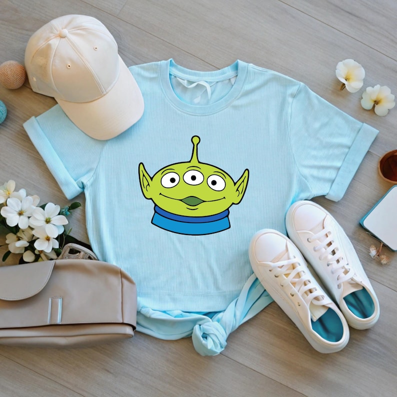 May include: Light blue t-shirt featuring a cartoon alien face with three eyes, green skin, and a blue collar. The shirt is paired with white sneakers and a white baseball cap. The shirt is made of a soft material.