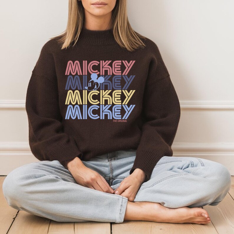 May include: A brown sweater featuring the word "MICKEY" stacked four times in different colors, with a Mickey Mouse silhouette and the text "THE ORIGINAL." The sweater has a mock neck and is paired with light blue jeans.