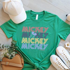 May include: Green t-shirt with the word "MICKEY" stacked four times in different colors. A Mickey Mouse silhouette is between the second and third "MICKEY" text. A white baseball cap, tan purse, and white sneakers are also in the image.