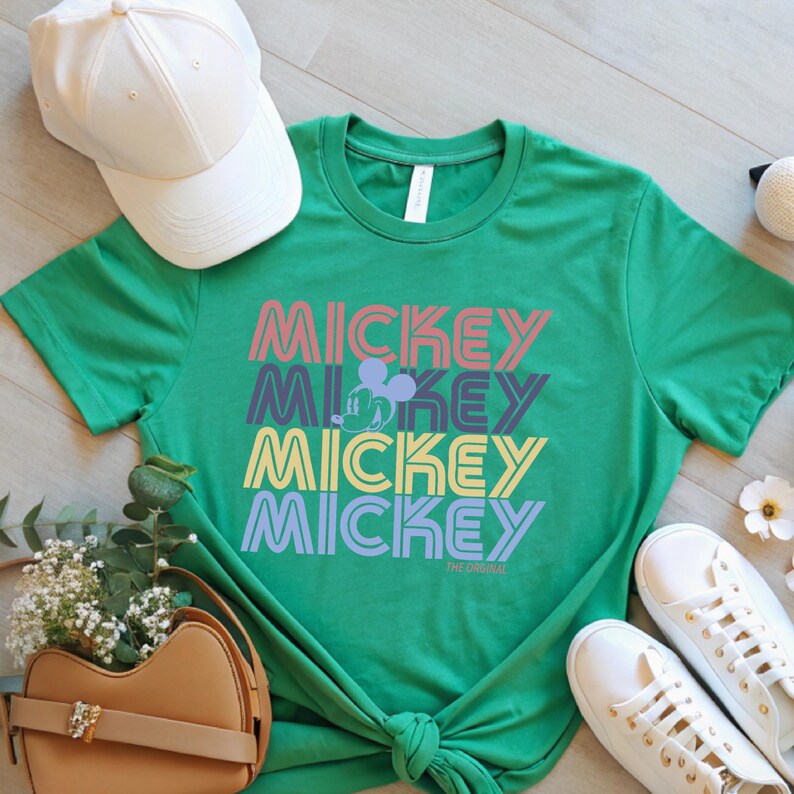 May include: Green t-shirt with the word "MICKEY" stacked four times in different colors. A Mickey Mouse silhouette is between the second and third "MICKEY" text. A white baseball cap, tan purse, and white sneakers are also in the image.